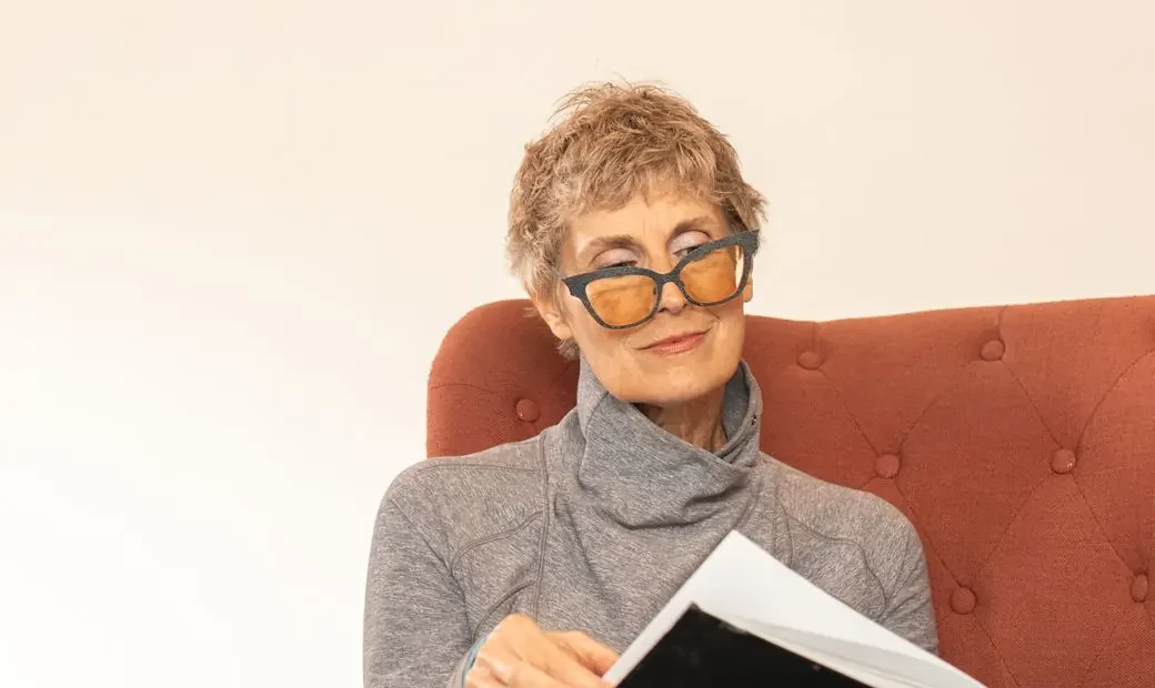 Une femme âgée aux lunettes ambrées sourit en lisant, installée dans un fauteuil.