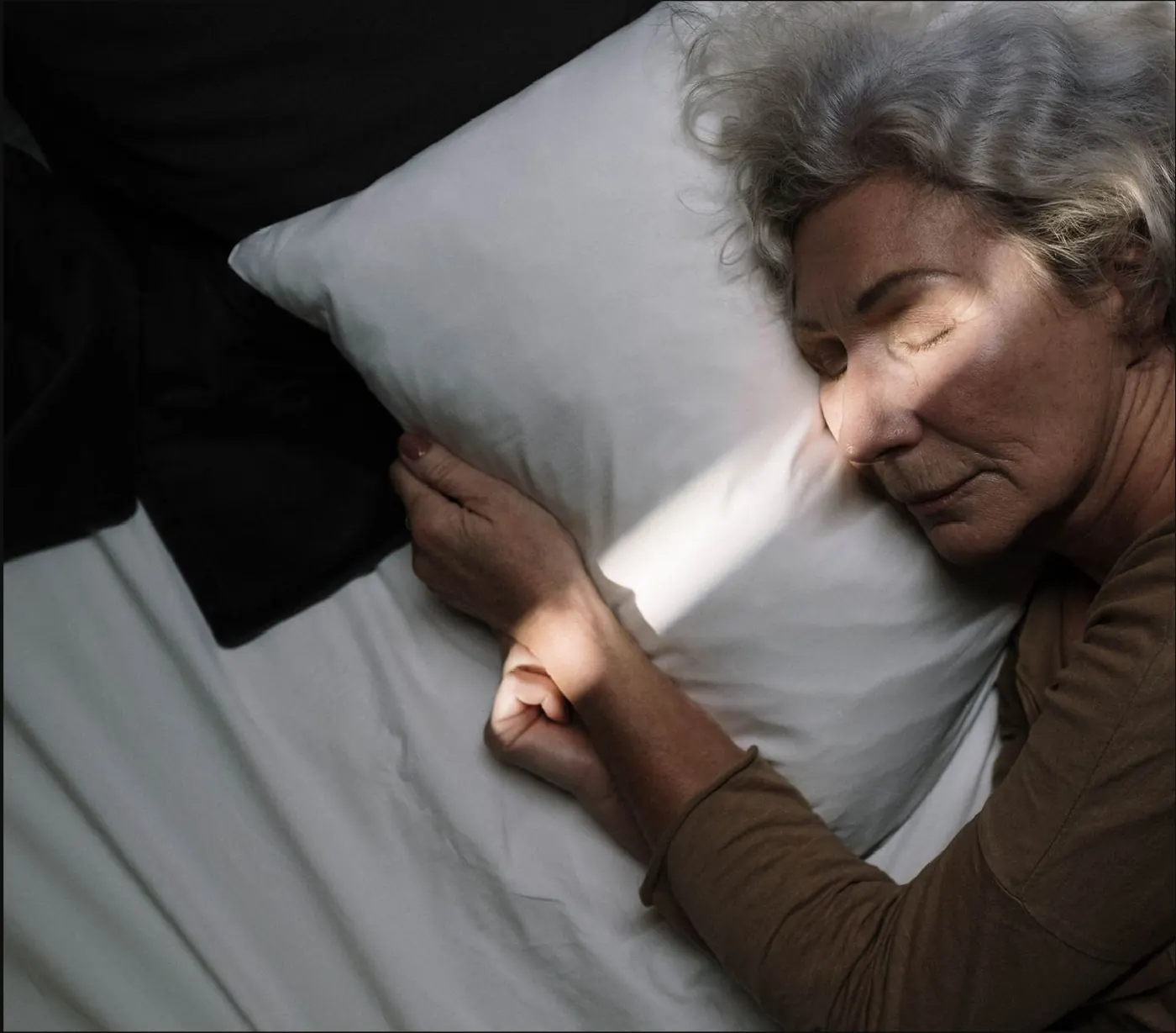 Une femme âgée repose, la tête sur un oreiller, dans la pénombre.
